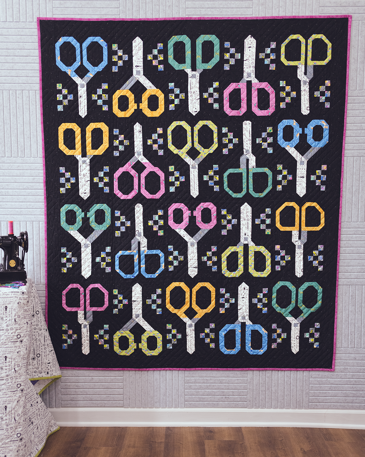 Quilt with colorful scissors pattern hanging on a wall in a room with sewing equipment.